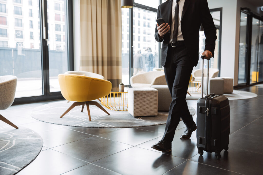 Cropped Photo Of Successful Businessman Wearing Suit Holding Smartphone And Walking With Suitcase In Hotel Lobby