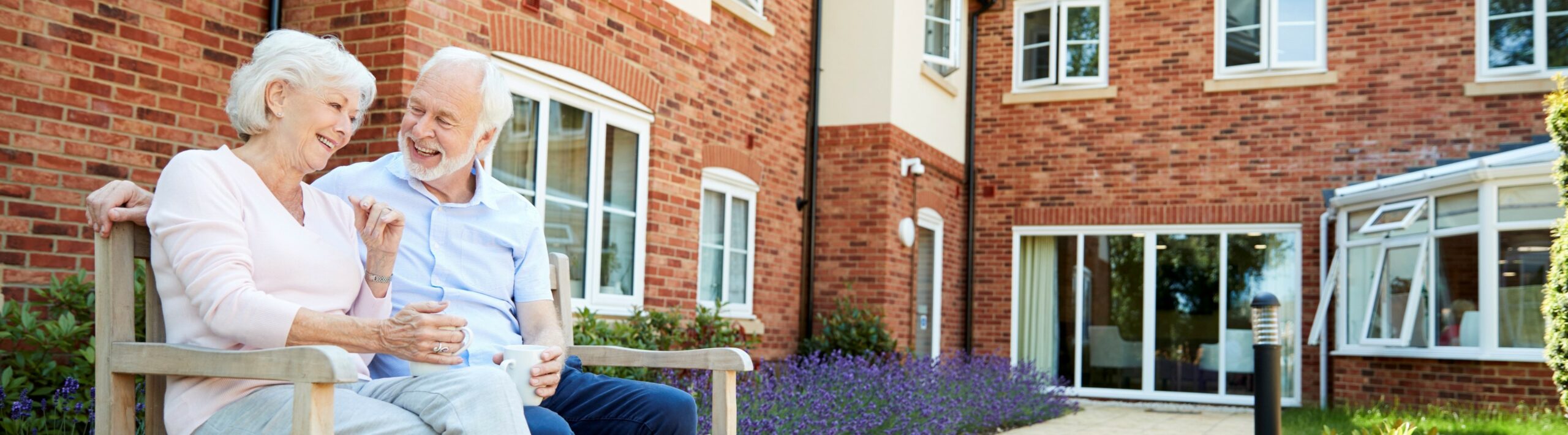 Retired Couple Sitting On Bench With Hot Drink In Assisted Living Facility